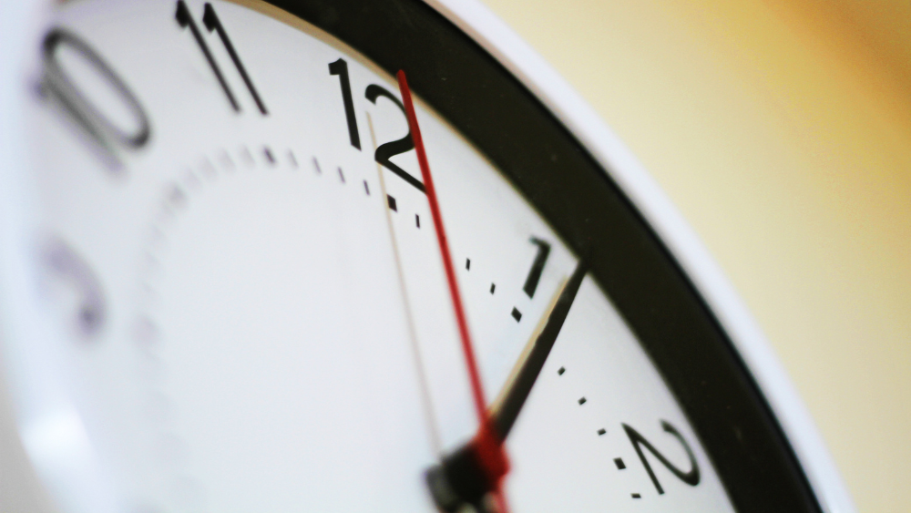 Nahaufnahme einer Wanduhr, die 12:02 Uhr zeigt. Der Sekundenzeiger bewegt sich auf die 12 zu.